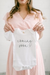 A person is holding a small white baby onesie with the words 'coming soon...' printed on it. The person appears to be in soft focus, wearing a light pink dress with a subtle pattern. The background is neutral, enhancing the focus on the onesie.