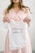 A person is holding a small white baby onesie with the words 'coming soon...' printed on it. The person appears to be in soft focus, wearing a light pink dress with a subtle pattern. The background is neutral, enhancing the focus on the onesie.