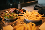 A rustic wooden table features three dishes: a bowl of fresh salad with leafy greens and a light dressing, a bowl overflowing with golden french fries, and a platter of grilled sausages accompanied by small roasted tomatoes and garnished with pickled onions. The scene has a cozy, homely feel with a background suggesting a casual dining environment.