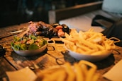 A rustic wooden table features three dishes: a bowl of fresh salad with leafy greens and a light dressing, a bowl overflowing with golden french fries, and a platter of grilled sausages accompanied by small roasted tomatoes and garnished with pickled onions. The scene has a cozy, homely feel with a background suggesting a casual dining environment.