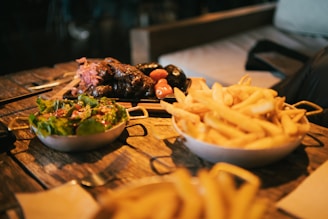 A rustic wooden table features three dishes: a bowl of fresh salad with leafy greens and a light dressing, a bowl overflowing with golden french fries, and a platter of grilled sausages accompanied by small roasted tomatoes and garnished with pickled onions. The scene has a cozy, homely feel with a background suggesting a casual dining environment.