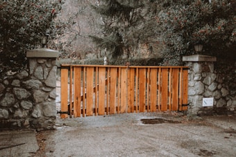 A wooden gate is placed between two stone pillars with foliage surrounding it. The gate is made of vertical wooden slats and is positioned on a gravel path. There are lamps on top of each pillar, and signs are visible next to one of the pillars.
