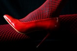A single red high heel shoe paired with black fishnet stockings that have red highlights. The background is dark, enhancing the vibrant red color of the shoe and stockings.
