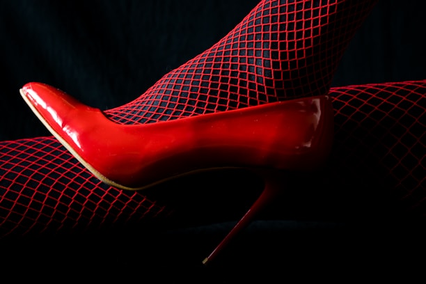 A single red high heel shoe paired with black fishnet stockings that have red highlights. The background is dark, enhancing the vibrant red color of the shoe and stockings.