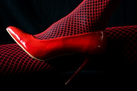 A single red high heel shoe paired with black fishnet stockings that have red highlights. The background is dark, enhancing the vibrant red color of the shoe and stockings.