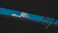 A cloud is seen through an opening in a building structure. The roof and beams frame a view of the sky, highlighting the fluffy white cloud against a blue background.