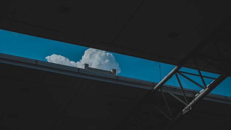 A cloud is seen through an opening in a building structure. The roof and beams frame a view of the sky, highlighting the fluffy white cloud against a blue background.