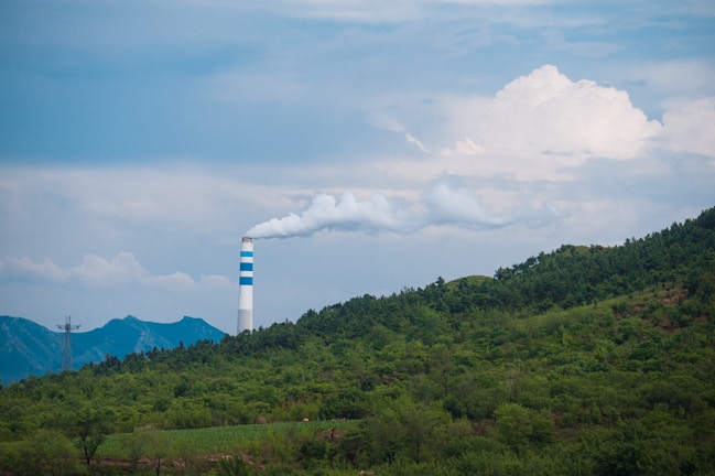 A tall smokestack with blue and white stripes releases smoke into a partly cloudy sky. It is surrounded by a lush green forest, with mountains visible in the background.