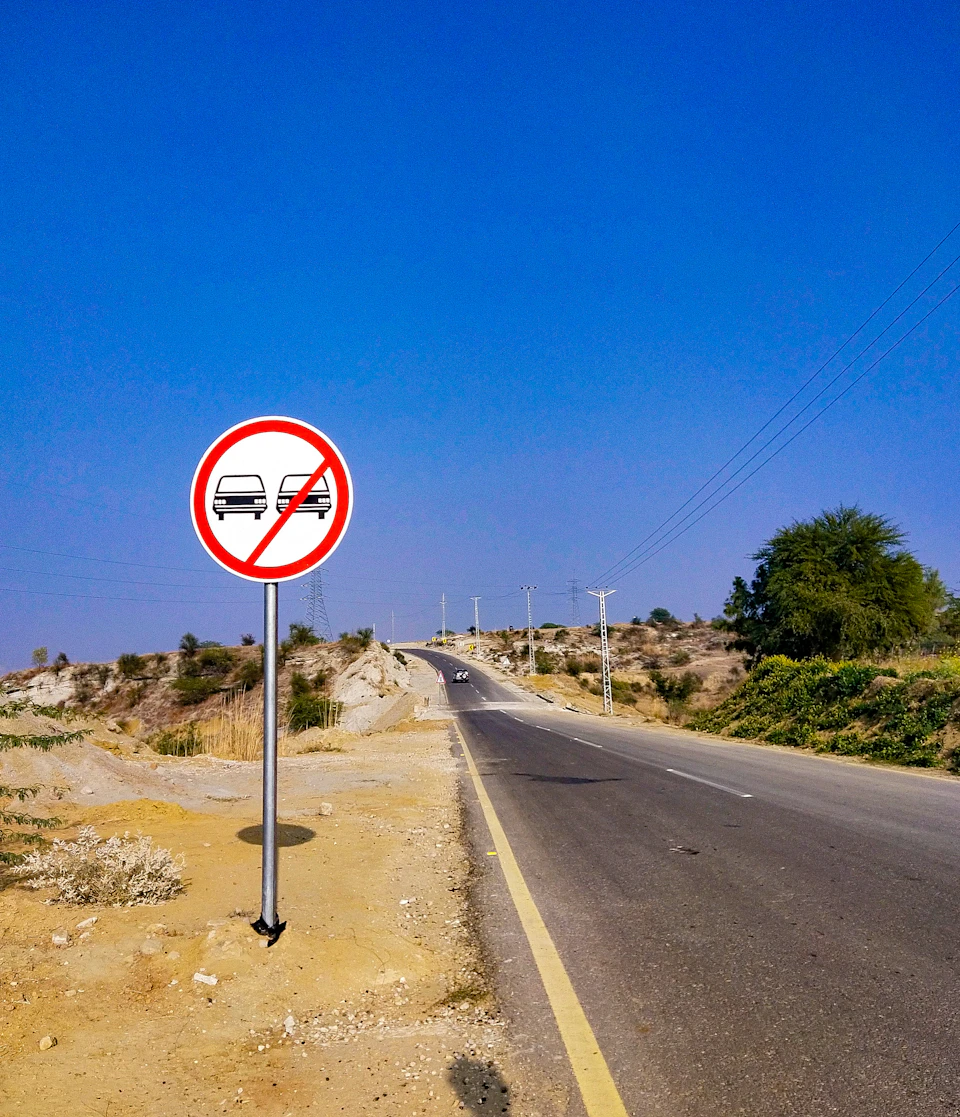 The image shows a two-lane road with a "No Parking" sign on the right side, indicating that parking is not allowed in the area. The road is surrounded by a desert landscape, and power lines can be seen in the distance. A few cars are parked along the roadside, but they appear to be following the rules of the no parking sign.
