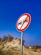 A circular road sign with a red border and a diagonal red line prohibits overtaking. Two car icons, one black and one red, are depicted inside the sign. The background features a clear blue sky and a natural landscape with a rocky hill and sparse vegetation.