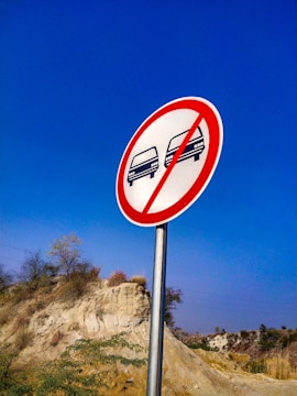 A circular road sign with a red border and a diagonal red line prohibits overtaking. Two car icons, one black and one red, are depicted inside the sign. The background features a clear blue sky and a natural landscape with a rocky hill and sparse vegetation.