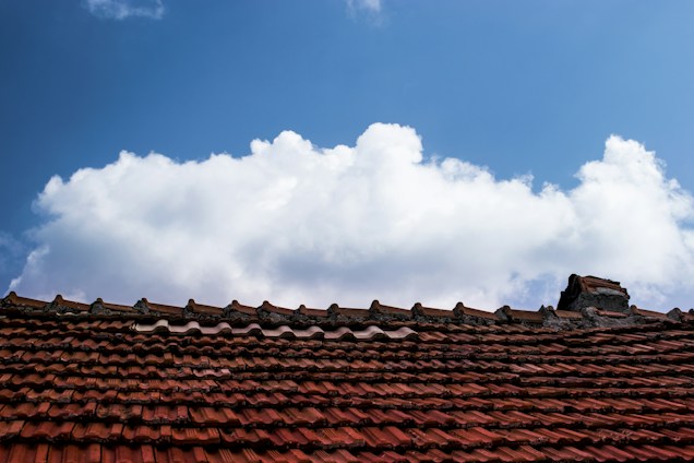 Red clay tiles compose the roof of a structure set against a vast, vibrant blue sky. Fluffy white clouds are scattered across the upper part of the image, creating a contrast with the solid lines of the roof.