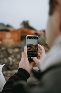 A person is holding a smartphone and taking a photo of a scene with the camera app open. The focus is on the phone's screen, which shows a landscape with some buildings or structures. The background is blurred, emphasizing the phone and hands.
