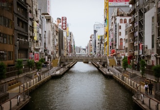 A canal lined with concrete pathways runs through a bustling urban area filled with tall buildings and vibrant billboards. Pedestrians can be seen walking along the paths, and there is a bridge crossing over the canal in the middle distance. The architecture is a mix of modern and traditional styles, with various signs and advertisements adding color to the scene.