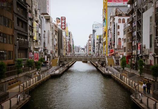 A canal lined with concrete pathways runs through a bustling urban area filled with tall buildings and vibrant billboards. Pedestrians can be seen walking along the paths, and there is a bridge crossing over the canal in the middle distance. The architecture is a mix of modern and traditional styles, with various signs and advertisements adding color to the scene.