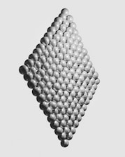 A geometric arrangement of metallic spheres forming a diamond-shaped three-dimensional structure. The spheres are uniformly spaced and appear highly polished, reflecting light subtly across their curving surfaces. The background is a plain, light gray, which highlights the structure's intricate pattern and symmetry.