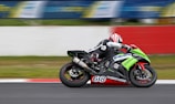 A motorcyclist wearing protective gear and a helmet is racing on a sportbike with bright green and black colors on a track. The motorcycle is marked with the number 618 and various brand logos. The background is blurred, indicating high speed, with a white and red barrier visible along the side of the track.