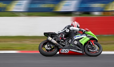 A motorcyclist wearing protective gear and a helmet is racing on a sportbike with bright green and black colors on a track. The motorcycle is marked with the number 618 and various brand logos. The background is blurred, indicating high speed, with a white and red barrier visible along the side of the track.