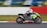 A motorcyclist wearing protective gear and a helmet is racing on a sportbike with bright green and black colors on a track. The motorcycle is marked with the number 618 and various brand logos. The background is blurred, indicating high speed, with a white and red barrier visible along the side of the track.