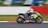A motorcyclist wearing protective gear and a helmet is racing on a sportbike with bright green and black colors on a track. The motorcycle is marked with the number 618 and various brand logos. The background is blurred, indicating high speed, with a white and red barrier visible along the side of the track.