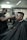 A barber is giving a haircut to a seated male client. The client is draped with a black haircutting cape, while the barber carefully uses clippers. The background includes multiple barber chairs and blurred light fixtures.