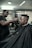 A barber is giving a haircut to a seated male client. The client is draped with a black haircutting cape, while the barber carefully uses clippers. The background includes multiple barber chairs and blurred light fixtures.