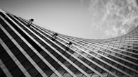 A curved, reflective skyscraper facade with several silhouetted workers suspended on ropes, engaged in cleaning or maintenance work. The contrast between the modern architecture and the cloudy sky creates a dramatic atmosphere.