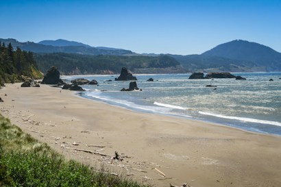 A serene coastal landscape features a wide sandy beach with scattered driftwood. Rocky formations jut out of the sea, and waves gently lap against the shore. The background is dominated by lush green hills and distant mountains under a bright blue sky.