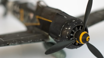 Close-up of a vintage World War II-era fighter plane model with a focus on the propeller and nose. The aircraft is painted in dark colors with yellow stripes detailing its body.