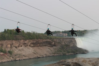 Three people wearing helmets are zip-lining across a large canyon with a waterfall in the background. The landscape features rocky cliffs and lush greenery. Mist from the waterfall creates a dramatic effect.