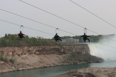 Three people wearing helmets are zip-lining across a large canyon with a waterfall in the background. The landscape features rocky cliffs and lush greenery. Mist from the waterfall creates a dramatic effect.