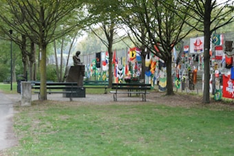 A peaceful park setting with a pathway lined by trees. A statue is seated on a bench, facing a colorful display of various flags and banners, which are attached to a wire fence. The grassy area is surrounded by trees, creating a serene atmosphere.