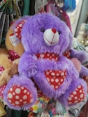 A fluffy purple teddy bear with a white and red polka-dotted heart on its chest. The bear has a friendly expression with round black eyes and a red nose. It features matching red and white polka-dotted ears and feet, and it is surrounded by various objects and possibly other toys in the background.
