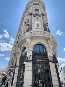 A grand and ornate building with intricate architectural details, featuring tall columns, decorative lamps, and sculptured elements. The facade displays signs for prominent luxury brands, located at a street corner under a clear blue sky.