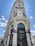 A grand and ornate building with intricate architectural details, featuring tall columns, decorative lamps, and sculptured elements. The facade displays signs for prominent luxury brands, located at a street corner under a clear blue sky.