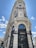 A grand and ornate building with intricate architectural details, featuring tall columns, decorative lamps, and sculptured elements. The facade displays signs for prominent luxury brands, located at a street corner under a clear blue sky.