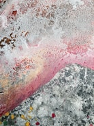 An abstract painting featuring a variety of textures and colors. The surface has a weathered appearance with a crackled pattern in off-white, covering parts of the canvas. Swathes of pink and red blend with earthy browns and gray tones, accented by splashes of yellow and black.