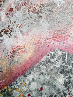 An abstract painting featuring a variety of textures and colors. The surface has a weathered appearance with a crackled pattern in off-white, covering parts of the canvas. Swathes of pink and red blend with earthy browns and gray tones, accented by splashes of yellow and black.