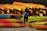 A person wearing a blue shirt with 'staff' written on it stands in front of a large stack of brightly colored kayaks. The kayaks are predominantly red, yellow, and orange, neatly arranged in several rows, with some green ones also present.