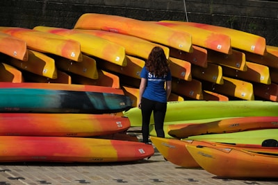 A person wearing a blue shirt with 'staff' written on it stands in front of a large stack of brightly colored kayaks. The kayaks are predominantly red, yellow, and orange, neatly arranged in several rows, with some green ones also present.