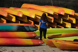 A person wearing a blue shirt with 'staff' written on it stands in front of a large stack of brightly colored kayaks. The kayaks are predominantly red, yellow, and orange, neatly arranged in several rows, with some green ones also present.