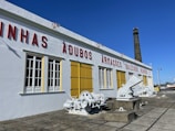 A historic industrial building with white walls and red lettering is situated under a clear blue sky. Yellow window shutters and large, white-painted machinery are visible outside. A tall brick chimney extends upward from the building, and there is a tiled stone pavement in front.