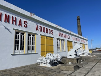 A historic industrial building with white walls and red lettering is situated under a clear blue sky. Yellow window shutters and large, white-painted machinery are visible outside. A tall brick chimney extends upward from the building, and there is a tiled stone pavement in front.