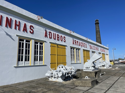 A historic industrial building with white walls and red lettering is situated under a clear blue sky. Yellow window shutters and large, white-painted machinery are visible outside. A tall brick chimney extends upward from the building, and there is a tiled stone pavement in front.