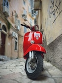 A red Vespa scooter is parked in a narrow, cobblestone alleyway. The old buildings surrounding the alley have rustic textures, graffiti on the walls, and window shutters, contributing to a vintage urban atmosphere.