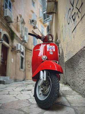 A red Vespa scooter is parked in a narrow, cobblestone alleyway. The old buildings surrounding the alley have rustic textures, graffiti on the walls, and window shutters, contributing to a vintage urban atmosphere.