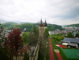A scenic view of a historical tower with pointed red roofs surrounded by lush greenery and distant mountains. In the foreground, there is a school sports complex with running tracks and a green sports field. The surrounding area is a mix of residential buildings and forested hills under a cloudy sky.