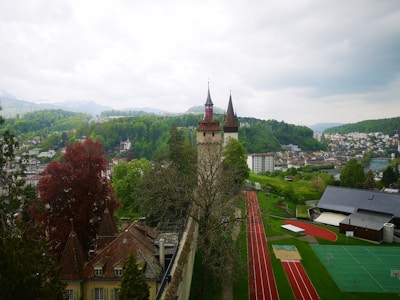 A scenic view of a historical tower with pointed red roofs surrounded by lush greenery and distant mountains. In the foreground, there is a school sports complex with running tracks and a green sports field. The surrounding area is a mix of residential buildings and forested hills under a cloudy sky.