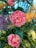 A vibrant and colorful bouquet featuring prominently pink roses, surrounded by purple and yellow flowers. The lush green foliage complements the blossoms, creating a lively and fresh floral arrangement.