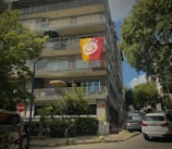 A multi-story residential building with a large flag displaying a sports emblem hanging from one of the balconies. The street is lined with trees, and several cars are parked alongside the building. A street sign and urban elements like a trash bin can be seen, creating a typical city neighborhood feel.